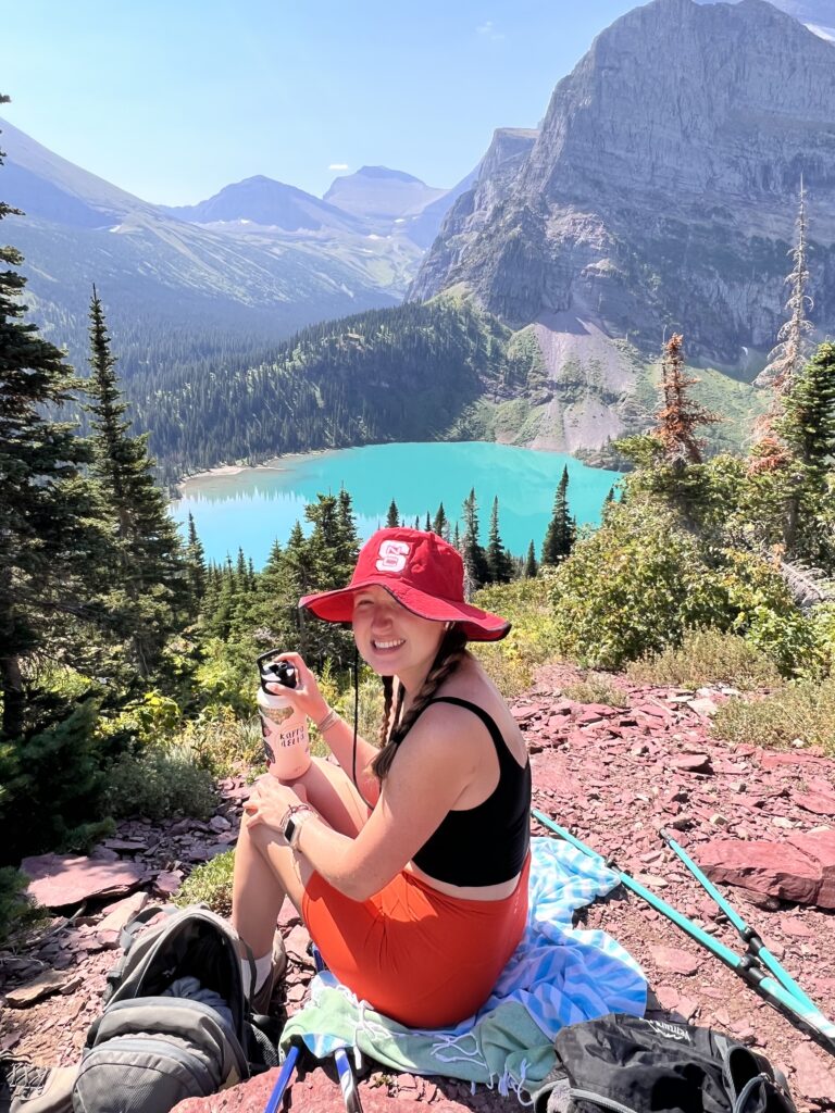 Hiker eating lunch on a rock overlooking a neon blue Grinnell Lake with mountains in the background